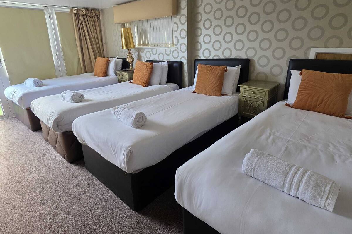 A hotel room with three beds, each with white linens and brown pillows, a bedside table with a lamp, and a patterned wallpaper.
