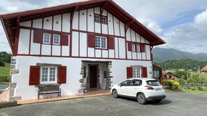 Una casa de dos pisos con un tejado inclinado, paredes blancas con detalles rojos y una base de piedra. Un coche blanco está estacionado adelante.