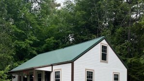 A white wooden house with a green roof, a porch, and a small garden with flowers and shrubs.