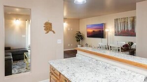 A modern kitchen with a marble countertop, a dining area with white chairs, and a painting of a sunset.