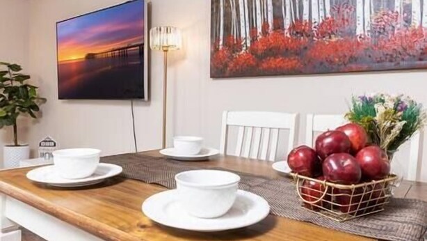 A dining area with a wooden table, white chairs, a painting of a sunset, and a basket of red apples.