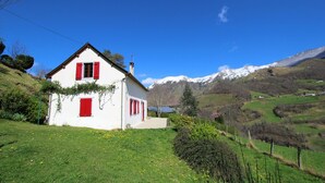A white house with red shutters and a small attached structure, surrounded by green fields and mountains in the background.
