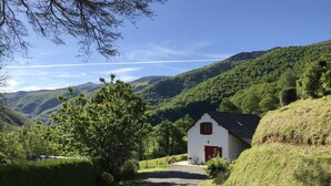 A rural setting with a white building featuring a red door and a sloped roof, surrounded by lush greenery and hills.