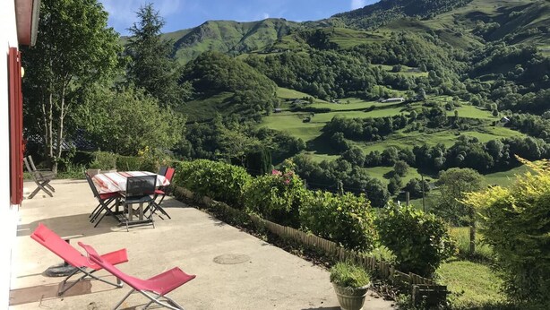 A patio with a table and chairs, overlooking a valley with green hills and a clear sky.