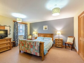 Ein Schlafzimmer mit einem Bett, zwei Nachttischen mit Lampen, einer Holzkommode, einem Fernseher und einem Fenster mit Vorhängen.