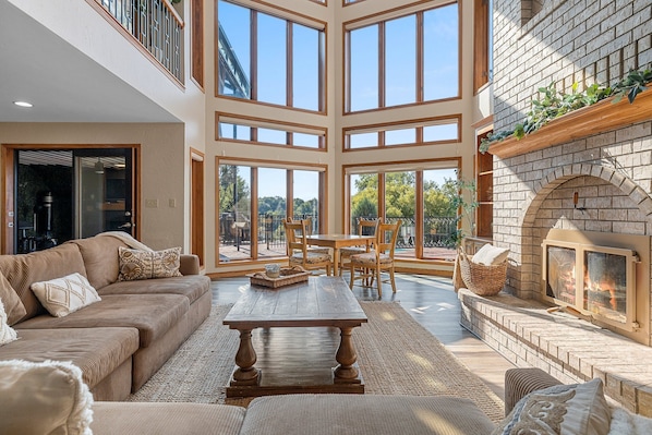 A spacious living area with a high ceiling, large windows, a fireplace, and a dining set.