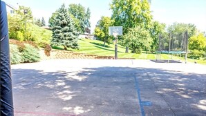 Una cancha de baloncesto al aire libre con un aro, rodeada de árboles y un muro de piedra.
