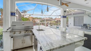 A spacious outdoor kitchen area with a built-in grill, a large marble countertop, and a view of a pool and surrounding landscape.