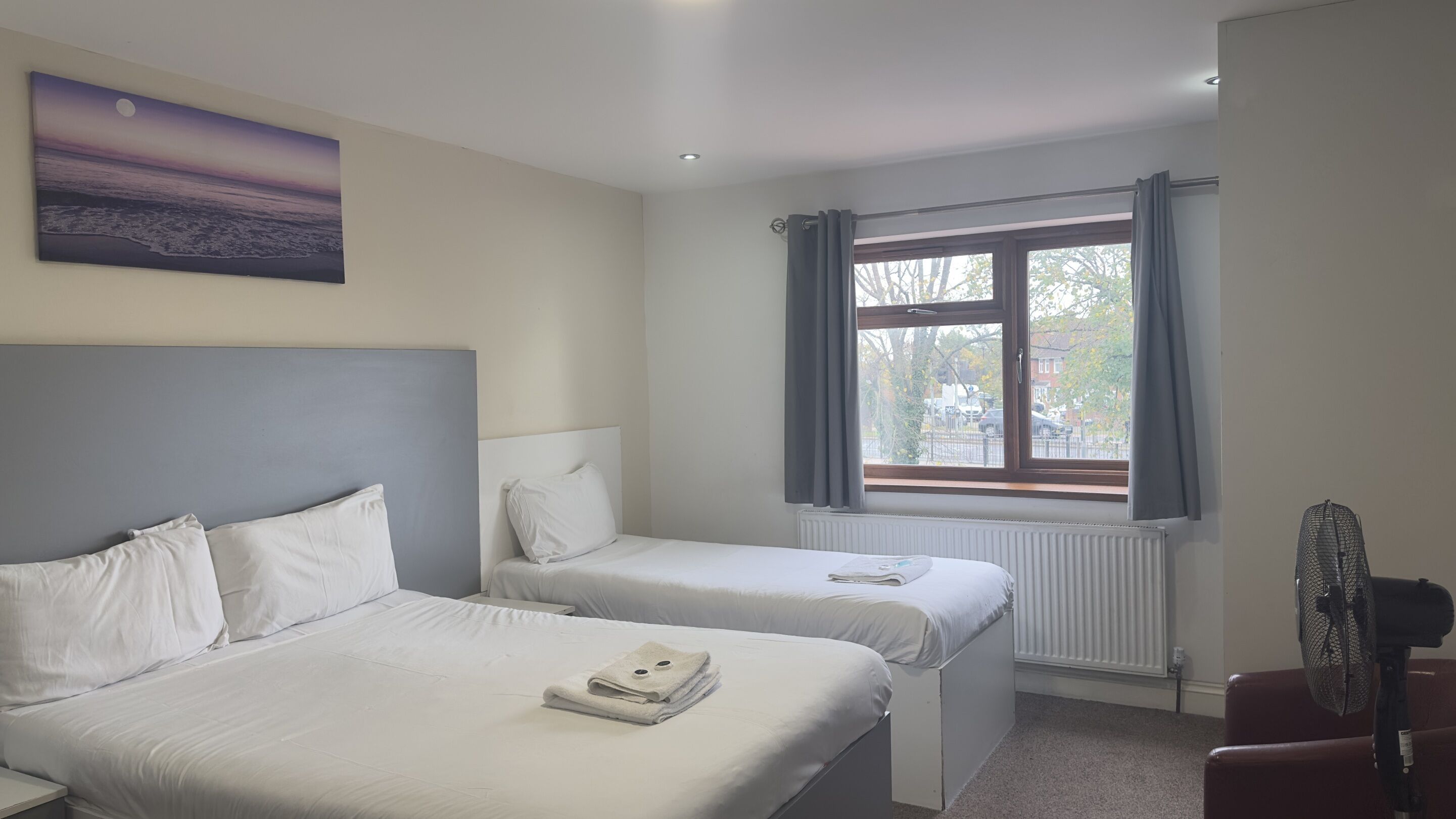 A hotel room with two single beds, a large window with curtains, a radiator, and a wall-mounted fan.