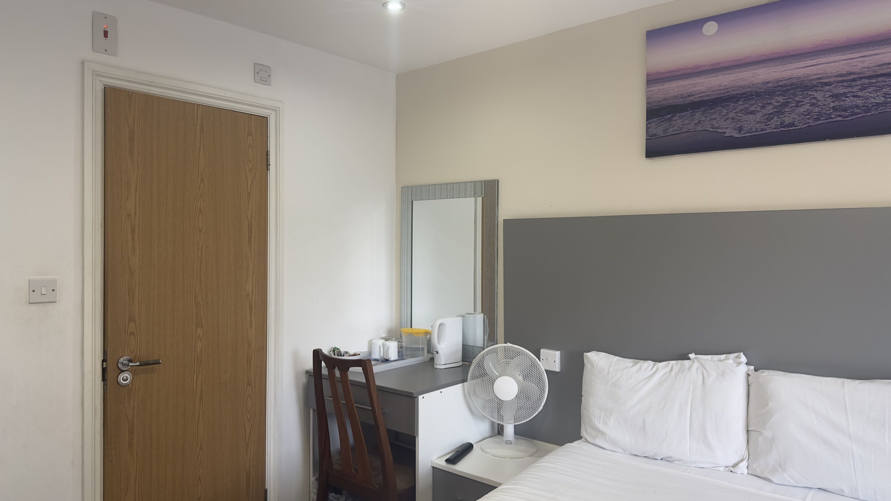 A hotel room with a wooden door, a desk with a chair, a bed with white bedding, a nightstand with a fan, and a painting of a sunset over the ocean on the wall.