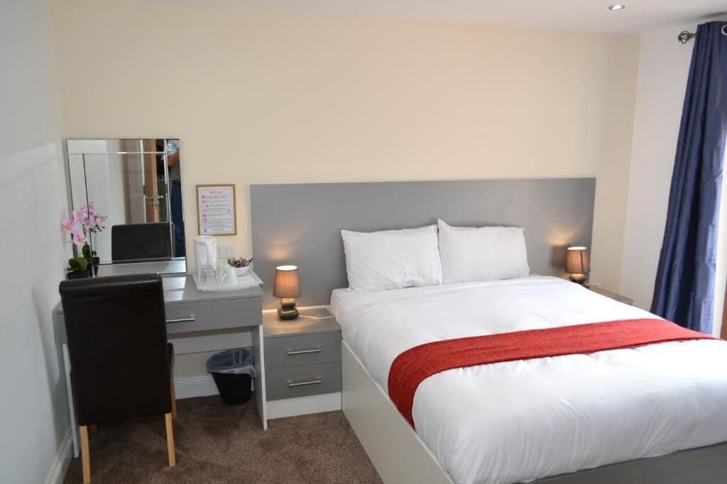 Una habitación de hotel con cama, mesita de noche, lámpara, silla, escritorio y espejo.