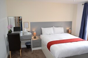 Una habitación de hotel con cama, mesita de noche, lámpara, silla, escritorio y espejo.