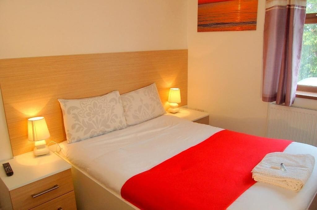 A neatly made bed with a red blanket, two bedside lamps, and a folded towel on the nightstand.