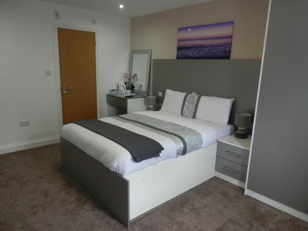 A bedroom with a bed, bedside table, and a nightstand. There is a door and a painting on the wall.