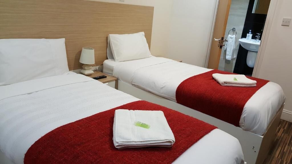 A hotel room with two beds, each with a red blanket and a folded white towel. There is a nightstand with a lamp and a small table between the beds. A bathroom is visible through an open door.