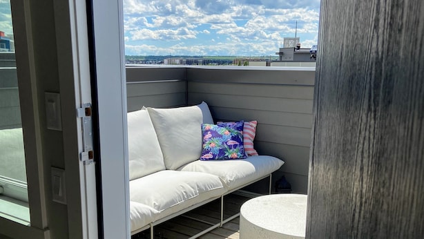 An open sliding door to a balcony with a white sofa, colorful pillows, and a round table. - 3 bedroom Townhouse Rooftop Patio & AC (Calgary)