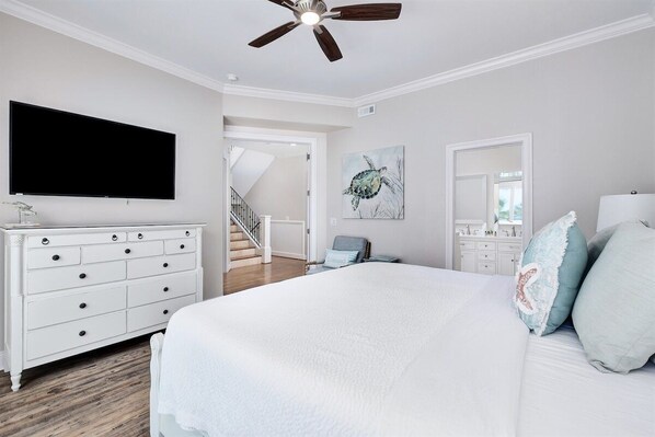 A bedroom with a large bed, a dresser, a flat-screen TV, a ceiling fan, and a staircase leading to another room.