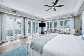 A spacious bedroom with a large bed, a ceiling fan, a television, and a balcony with a view of the ocean.