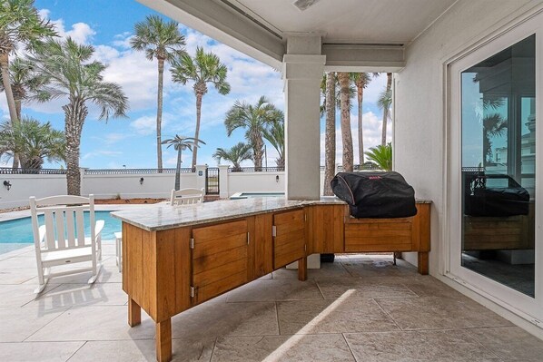 A covered outdoor area with a wooden bar and seating, overlooking a pool and palm trees.
