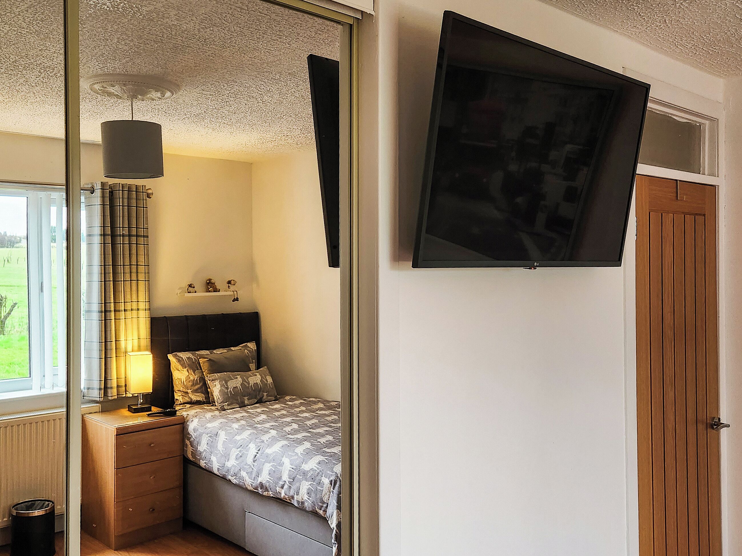 A bedroom with a bed, bedside table, lamp, and a TV mounted on the wall.