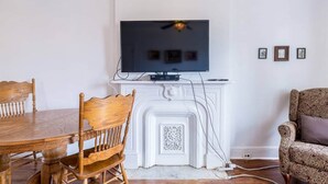Ein Essbereich mit einem Holztisch und Stühlen, einem weißen Kamin mit darüber installiertem Fernseher und einem gemusterten Sessel.