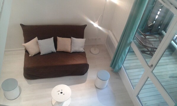 Un sofá marrón con cojines blancos en una habitación con piso blanco, una mesita pequeña blanca y una ventana con vista a una escalera.