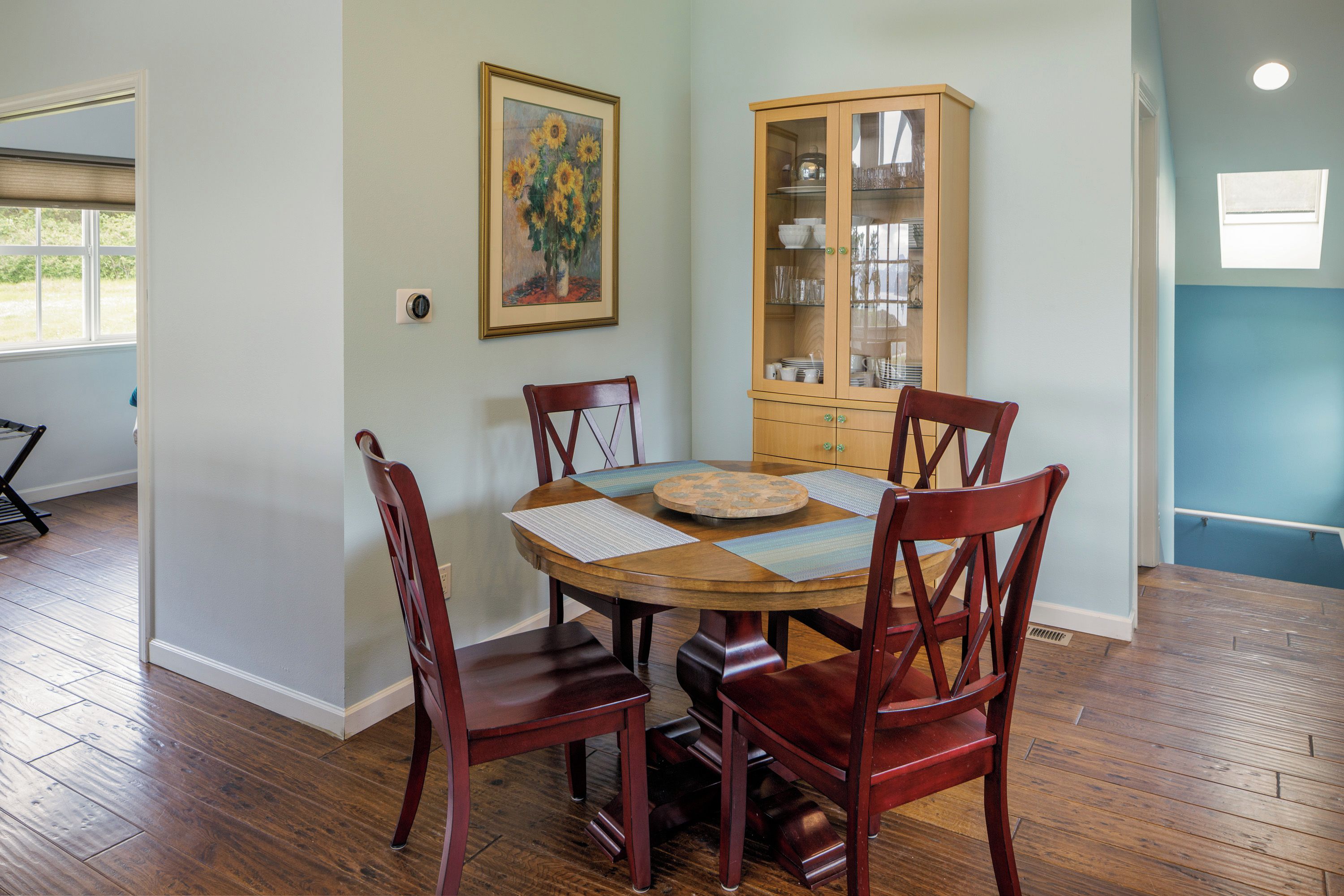 Un comedor con una mesa redonda de madera, cuatro sillas de madera, un mueble de madera con puertas de cristal y un cuadro enmarcado en la pared.