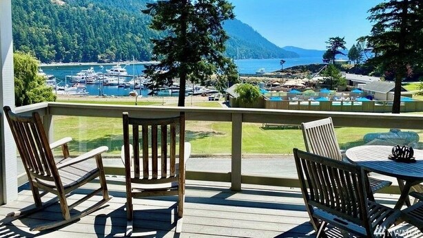 Un espace extérieur abrité, doté de chaises et d’une table en bois, qui offre une vue sur une marina avec des bateaux et un paysage montagneux.
