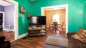 A living room with a television on a wooden stand, a dining table with chairs, and a couch with a patterned throw.