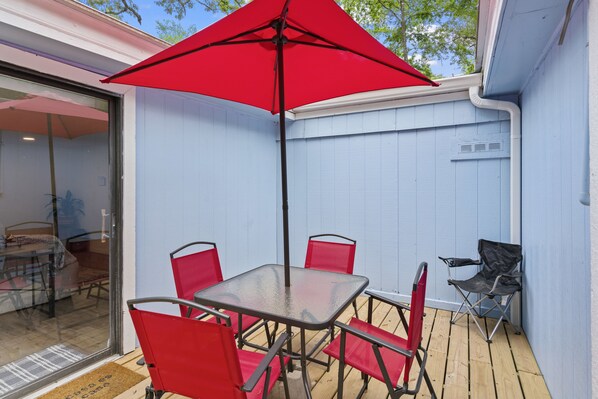 A patio with a glass door leading to a dining area, a table with four red chairs, and a red umbrella. - Magnolia House w/ 4 Beds 2 Baths on 1.5 Acres ⭐ (Magnolia)