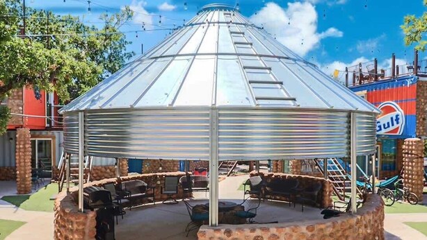 Um gazebo com cobertura metálica e base de pedra, rodeado por área de estar externa e churrasqueiras.