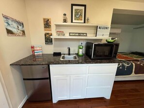 A compact kitchen with a built-in refrigerator, a microwave, and a sink. There are decorative items on the shelves, including a welcome sign and a framed picture. - HT Innhabit rooftop view-Hike Wichita Mtns-Slps4 (Medicine Park)