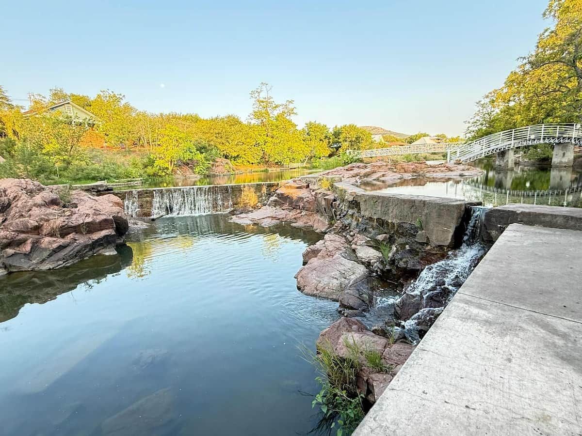 A tranquil outdoor setting with a small waterfall, a concrete pathway, and a metal bridge over a body of water.
