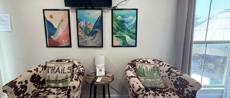 A room with two cowhide-patterned armchairs, a small round table with a lamp, and three framed artworks on the wall.