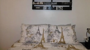 A bedroom with a bed featuring Eiffel Tower prints on the pillows, a wall-mounted photo collage, and a nightstand with a lamp.