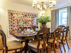A dining area with a wooden table, six chairs, a chandelier, a floral wall decoration, and a vase with green foliage.