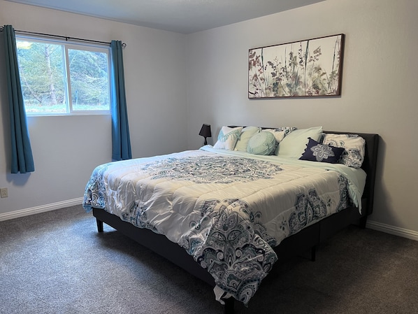 A bedroom with a large bed, a window with curtains, a wall-mounted artwork, and a carpeted floor.