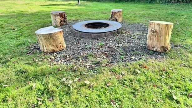 En rundad eldstad omgiven av fyra trädstampar, placerad på en gräsyta med en hög med träflis i mitten.