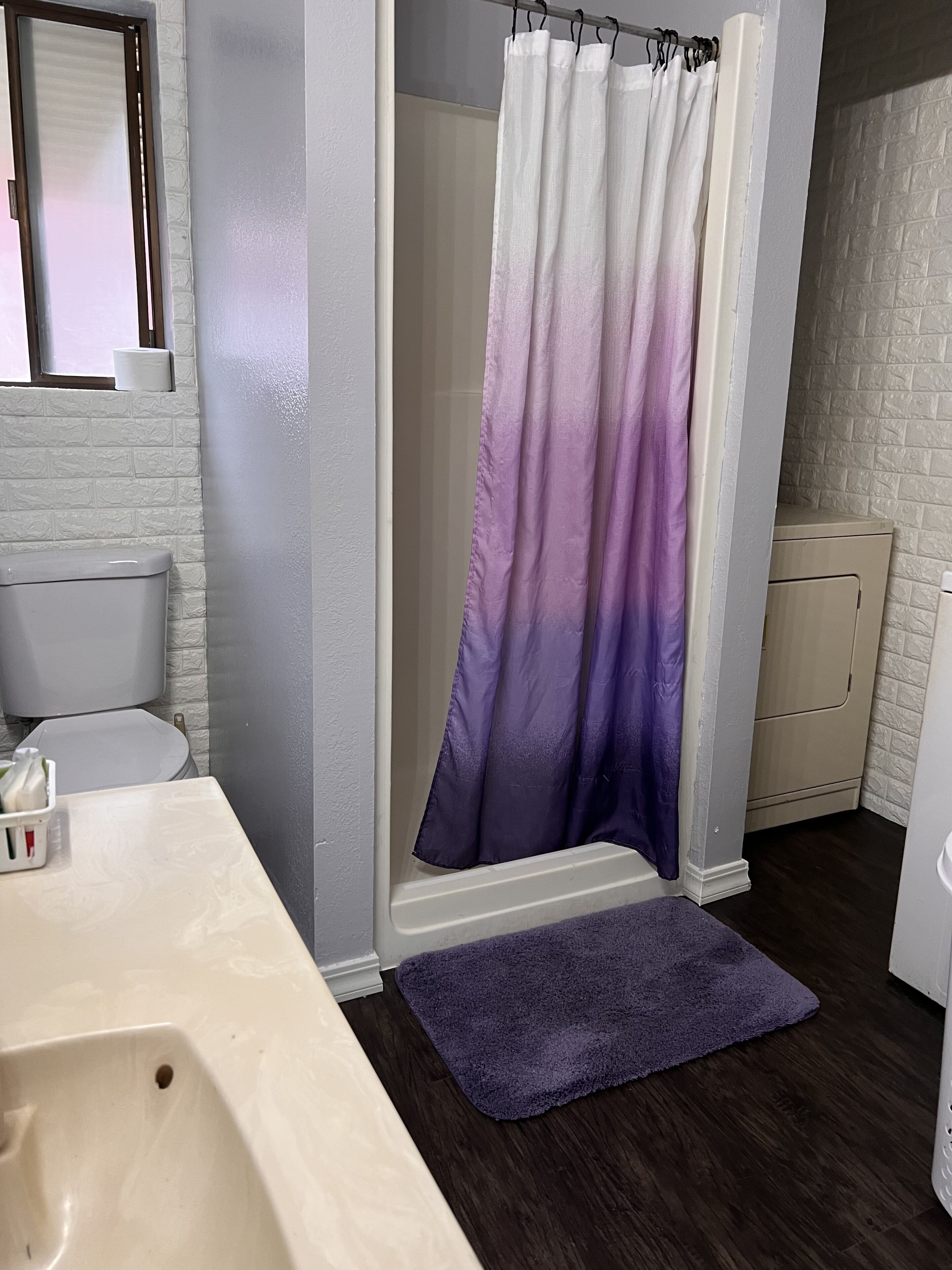 A bathroom with a white toilet, a white sink, a purple shower curtain, and a purple bath mat.