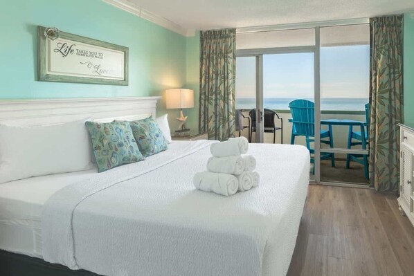 Een hotelkamer met een bed, uitzicht op zee, een balkon met stoelen en een citaat in een lijst op de muur.