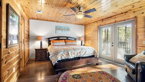 A wooden cabin bedroom with a bed, nightstands, a ceiling fan, and a large window.