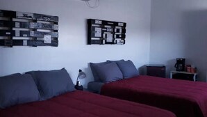 Ein Zimmer mit zwei Betten, jeweils mit roter und grauer Bettwäsche, einem kleinen Tisch mit einer Lampe sowie einer Wandtafel.