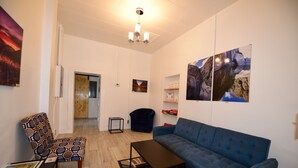 Una sala de estar moderna con un sofá azul, un sillón estampado y una pequeña mesa. Hay una puerta que conduce a otra habitación y un estante en la pared con libros.