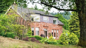 A stone house with a pitched roof, surrounded by lush greenery and trees.
