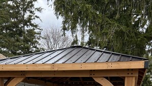 Eine überdachte Terrasse im Freien mit einer Holz-Pergola, einem grauen Ecksofa mit gestreiften Kissen und einem weißen Schaukelstuhl.