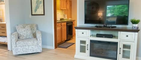A living room with a TV, a white cabinet with glass doors, a patterned armchair, and a kitchen in the background.