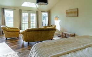 Une chambre spacieuse avec un grand lit, deux fauteuils, une lampe sur pied et une vue sur l’extérieur grâce à de grandes fenêtres.