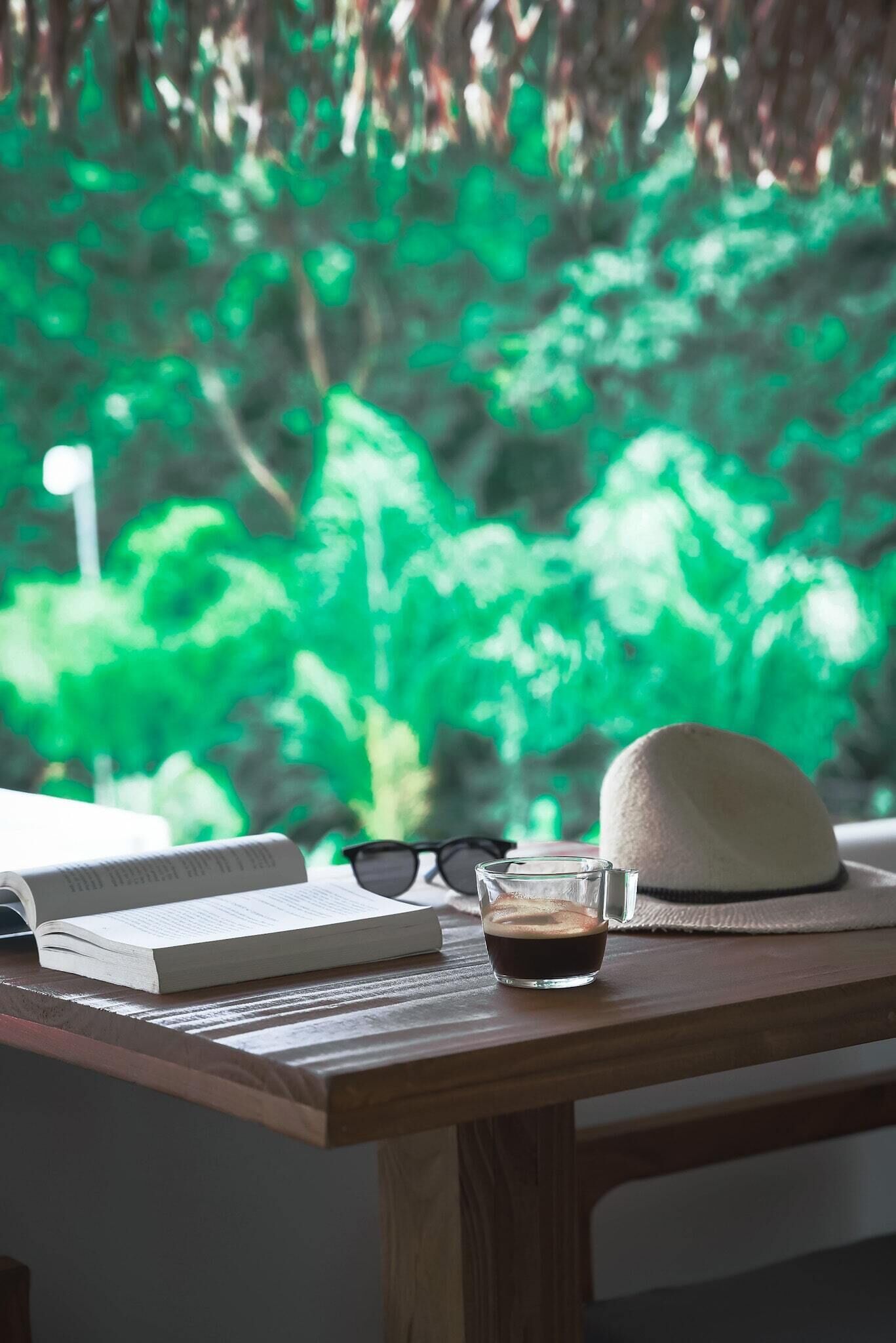 A wooden table with a book, a pair of glasses, a glass of coffee, and a hat.