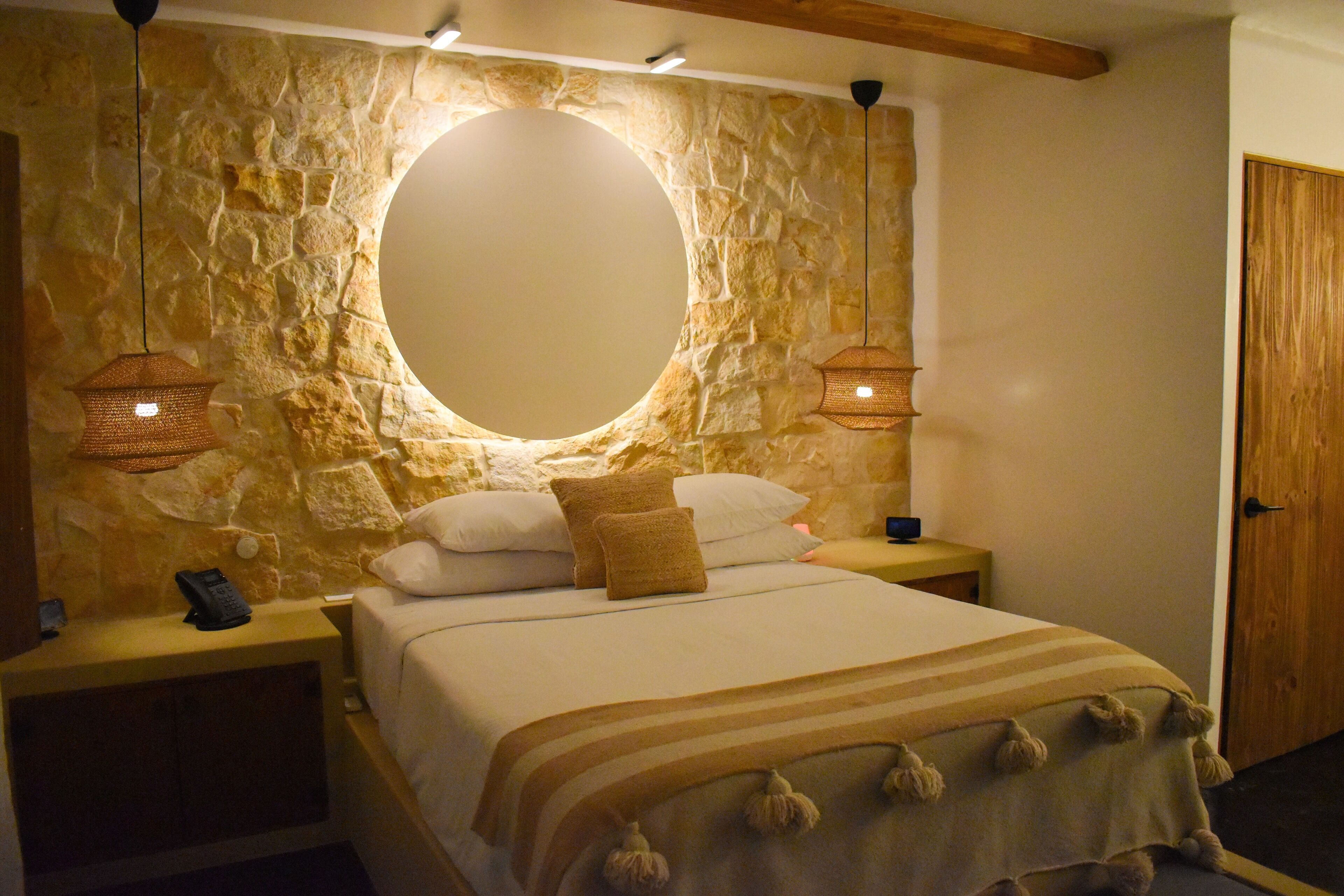 A bedroom with a stone wall, a round mirror, two bedside tables with lamps, a bed with a striped blanket, and a wooden door.