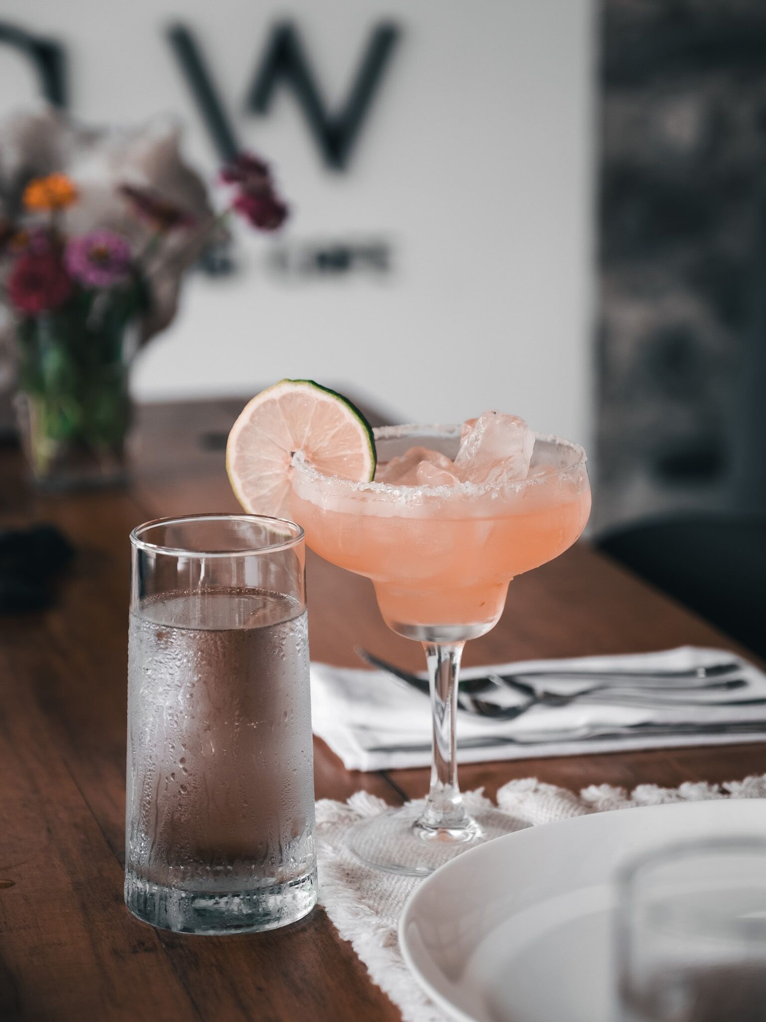 Un verre de cocktail rose, garni d’une tranche de citron vert et de glaçons, et un grand verre d’eau sur une table en bois.
